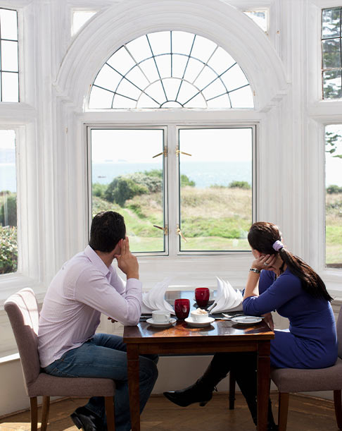 Man and woman seated at a dining table inside a bright bay window nook with arched transom, looking out at seaside landscape