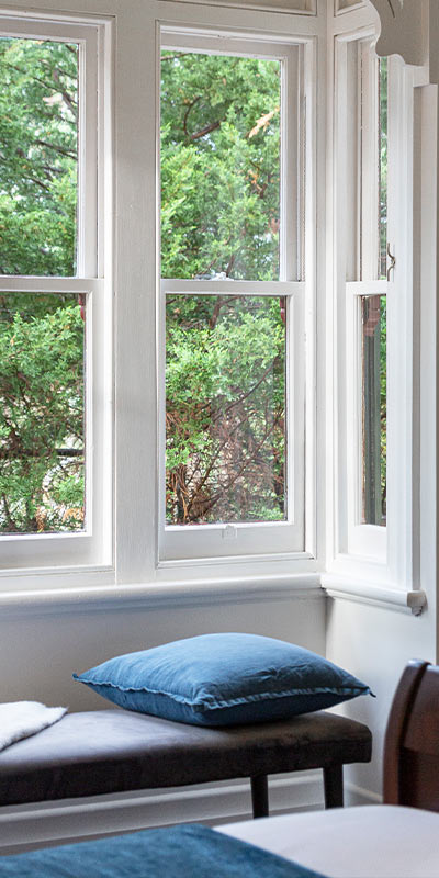 Interior bench topped with pillow beneath multi-lite bow windows, overlooking dense garden foliage